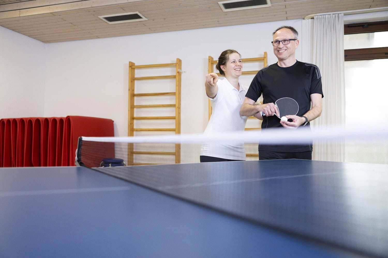 Therapeutin und Mann spielen Tischtennis in einer Sporthalle mit Sprossenwand