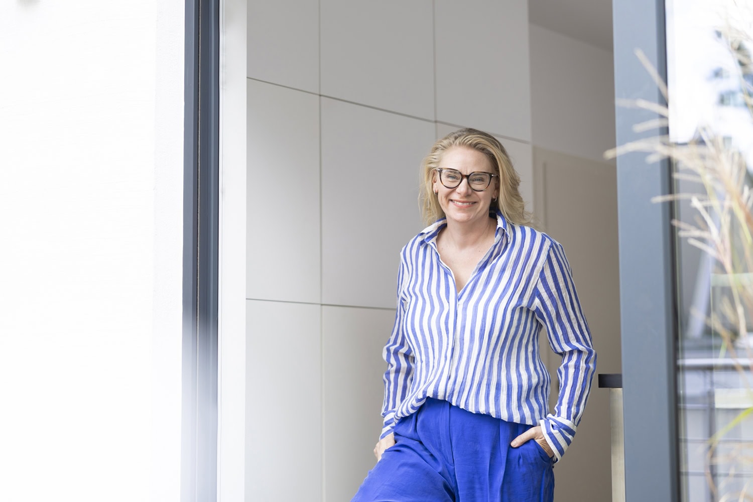 Lächelnde Businessfotografin Claudia Masur aus Leipzig in blau-weiß gestreifter Bluse vor modernem Büroflur – spezialisiert auf Unternehmensfotografie, Employer-Branding und Mitarbeiterporträts.