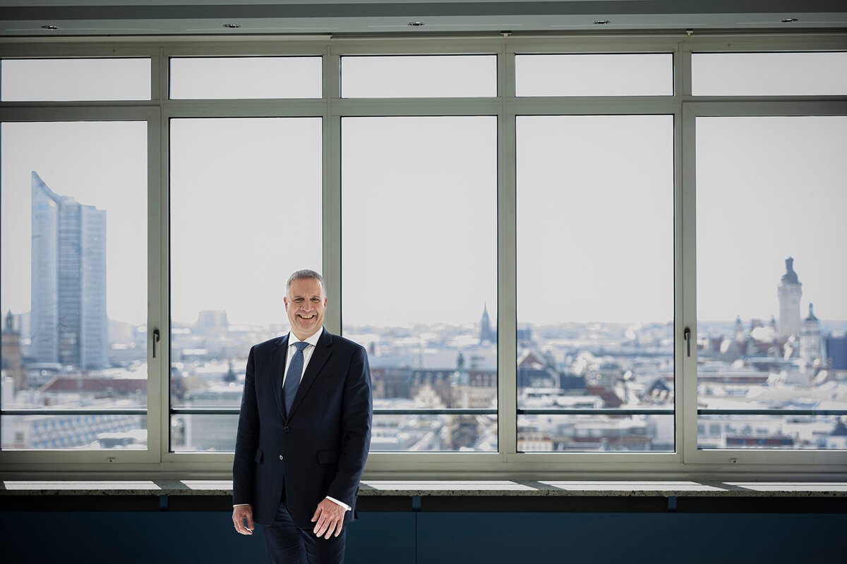 Ein Mitarbeiter der Stadtsparkasse Leipzig posieren für ein professionelles Businessportrait in einem modernen Konferenzraum mit urbanem Ausblick. Sie trägt elegante Kleidung und verkörpert Kompetenz, Seriosität und Leadership.