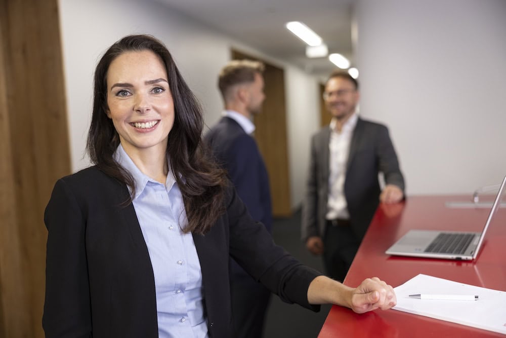 Lächelnde Mitarbeiterin am Empfangstresen, Kollegen im Hintergrund – Corporate Fotografie Leipzig, Claudia Masur Photography