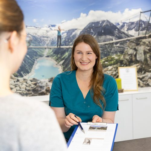 Mitarbeiterin in petrolfarbener Praxiskleidung vor der Wand mit Bergpanorama spricht mit einer Patientin am Empfang – Zahnarztpraxis Dr. Höhne, Leipzig